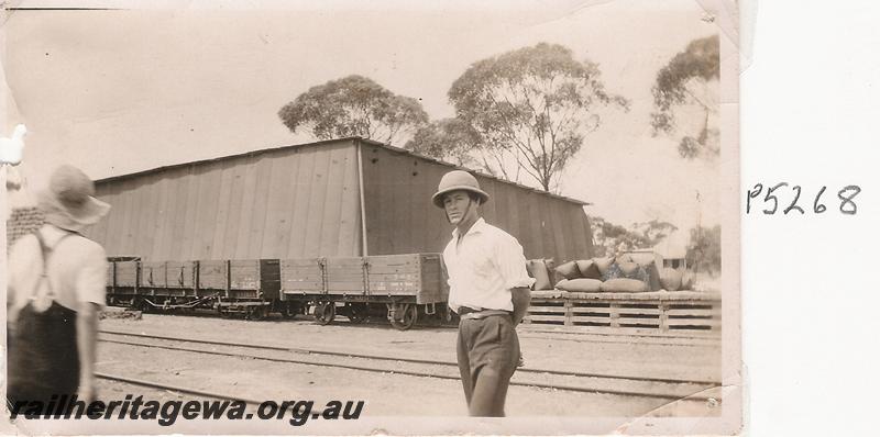 P05268
Station staff member wearing a pith helmet type hat, R class 1825 bogie open wagon and G class 2611 four wheel open wagon in background, large storage shed, location unknown, possibly on the CM line.
