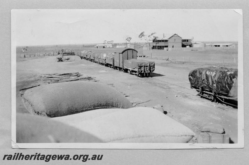 P08976
Tarpaulin covered wagons in siding, Mukinbudin, WLB line, station buildings in background, station nameboard with white lettering on a black background, hotel in the far background.
