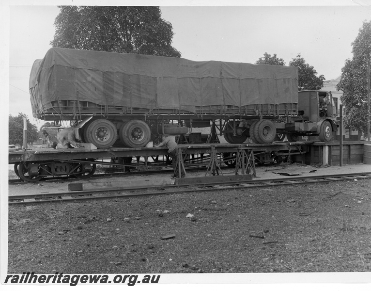 P10819
QM class flat bed wagon, with semi trailer partially on it and adjacent platform, workers placing chocks behind and in front of truck wheels, side view
