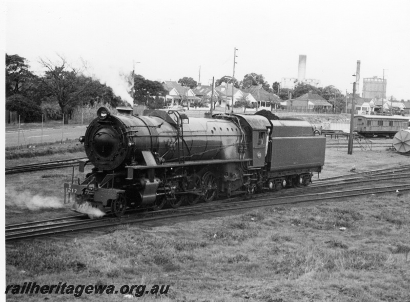 P17147
V class 1217 steam locomotive at eastern end of East Perth loco showing front/side view. JG fuel tank on ground at rear, workman's van at rear background and gasometer in rear far background.
