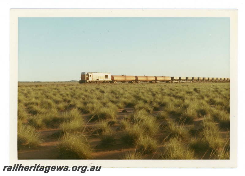 P18938
Goldsworthy Mining (GML) A class locomotive hauls an empty train across the spinifex plains between Port Hedland and Goldsworthy. Behind the locomotive is 3 water tank cars.
