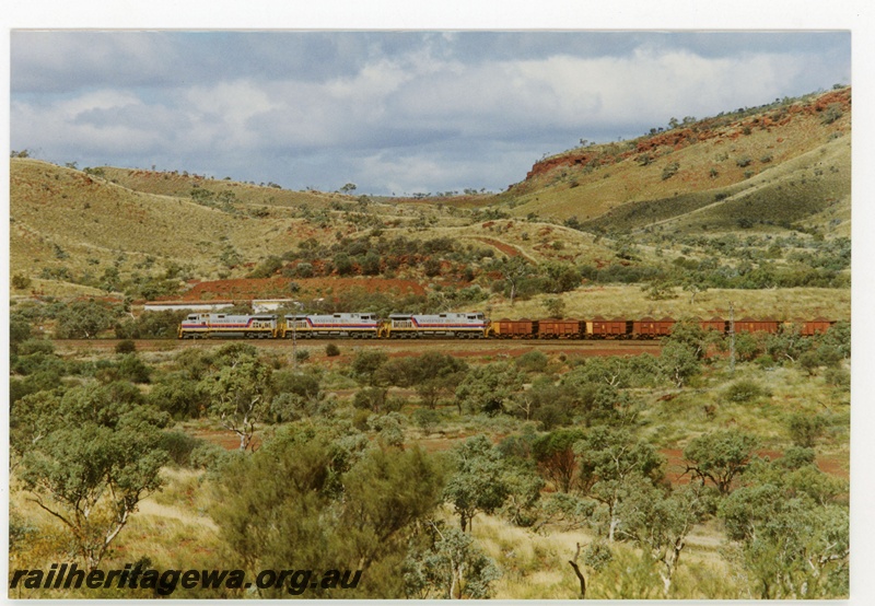 P18966
Hamersley Iron (HI) C44-9W class 7086, 7080, 7081 departing Tom Price with a loaded iron ore train. Side view of locomotives and several ore cars, with mountain range in background.
