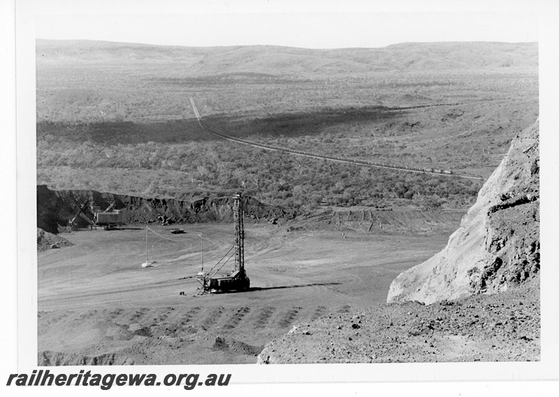 P19235
Mount Newman Mining (MNM) distant view of empty train arriving Newman with drill blast hole pattern in foreground.
