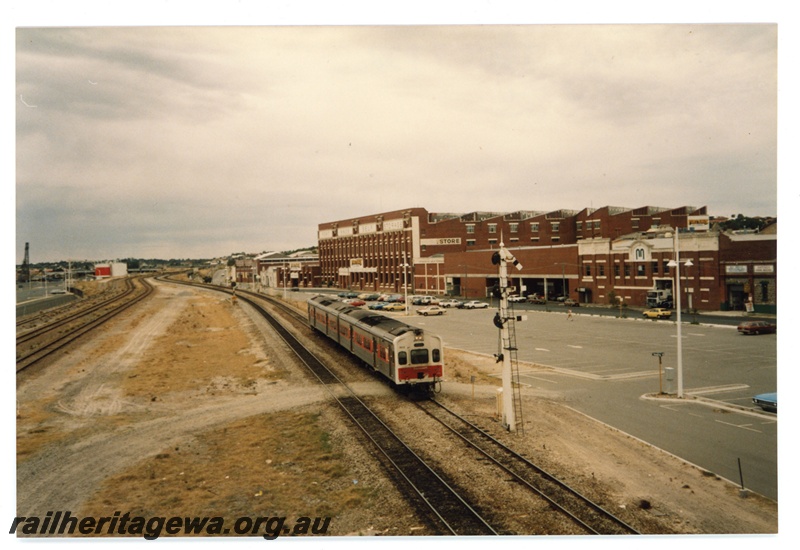 P19270
View of northern approach to Fremantle station, two car Transperth DMU travelling south, signal, wool stores, rail bridge over Swan River in background, side and front view of train, taken from pedestrian overpass
