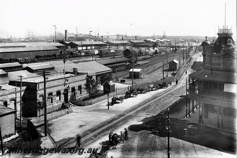 P21480
Original Fremantle railway station, station building, goods yard, warehouses, ship funnels and masts, overhead footbridge, horse drawn carts, His Majesty's hotel building, trackside sheds, tracks, Mouat Street, Fremantle, ER line, view from elevated position, c1905
