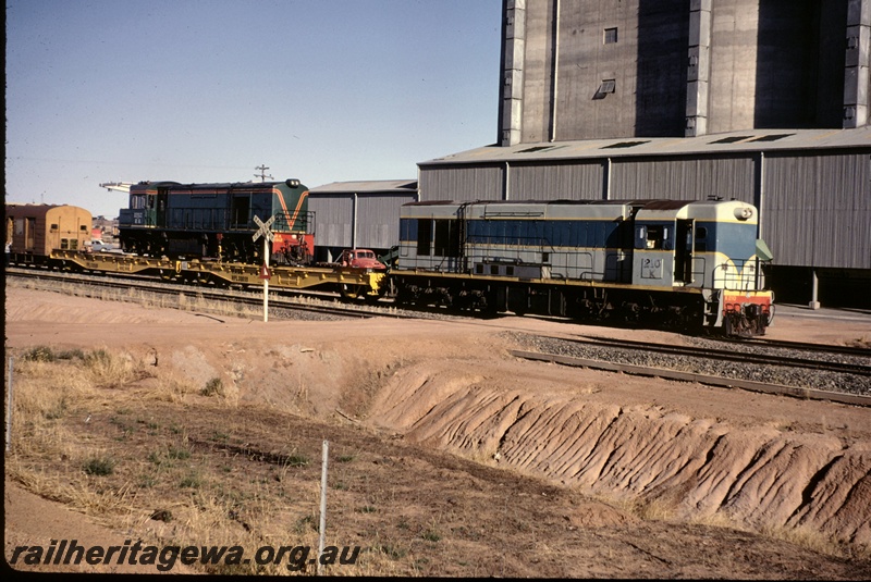 T05144
K class 210, on goods train including WFL class flat wagon 30059 and WFL class 30060 with RA class 1912 on board, and van, road crossing, wheat silos, West Merredin, EGR line
