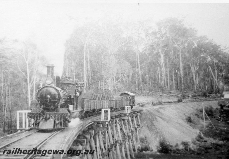 P02876
G class steam locomotive hauling a timber train including a brakevan across a large trestle bridge near Deanmill, three men on the footplate and many people on the brakevan, front and side view.
