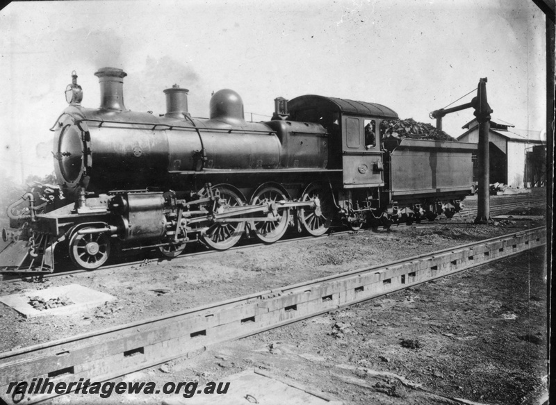 P03251
E class steam locomotive taking on water at a water column, front and side view, single stall loco shed, drip trays for water columns at each end of the engine pit.
