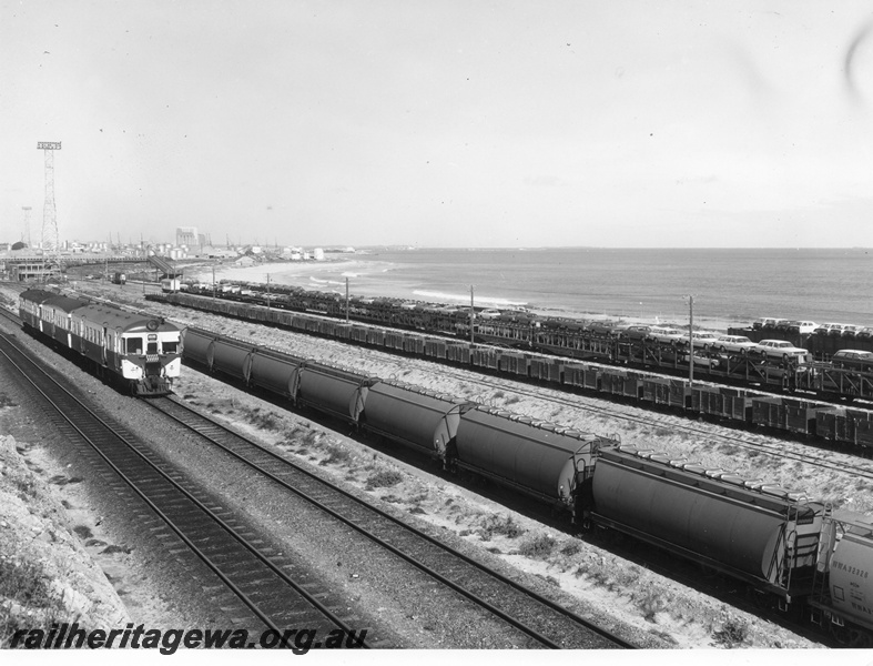 P10412
ADG/ADA/ADG class railcar set travelling east passing Leighton Marshalling Yard with standard gauge wheat hoppers in foreground. Standard gauge car carriers in background and narrow gauge single wagons in mid foreground. At left is Leighton Yard building and footbridge in distance at left as are light tower and fuel storage tanks. 
