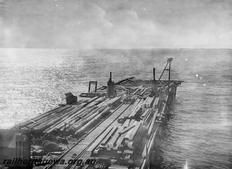 P10429
3 of 3 photos of construction of the Bunbury Jetty. Minimal sign of any safety equipment. Crane at right background, timber decking in foreground and piles on left of completed decking. Rail track down centre of area and small trolley at end.
