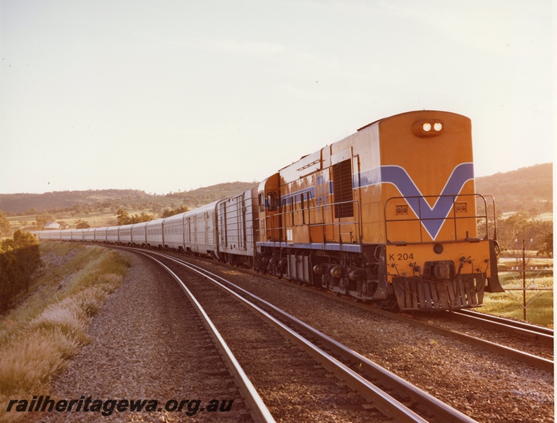 P10491
K class 204, in Westrail orange with blue and white stripe, heading passenger train of stainless steel carriages, in rural setting on dual gauge track, side and front view
