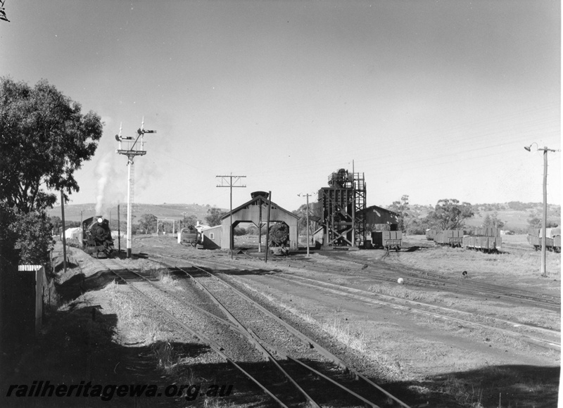 P15104
An unidentified W class steam locomotive arriving York from the Quairading Branch line. YB line. An unidentified X class diesel locomotive and V class steam locomotive at the loco depot together with GH class coal wagons at the coal stage and other wagons in the yard. Note the semaphore signal, yard lights and telephone pole.
