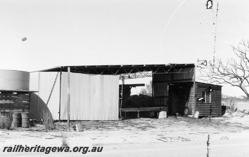 P15174
2 of three views of the sleeper mill located trackside at Narrikup, c1970/71, GSR line, roadside view of the mill shed and weatherboard office with a water tank on a stand at the left hand end
