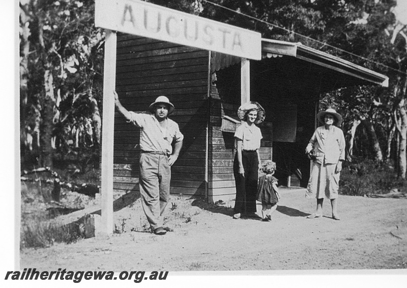 P15809
Station nameboard, shelter shed, Augusta, BB line, three adults and a small child in front of the nameboard
