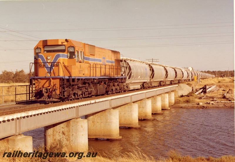 P15895
N class 1877, in Westrail orange with blue and white stripe, on alumina train, crossing low concrete and steel bridge over river, front and side view
