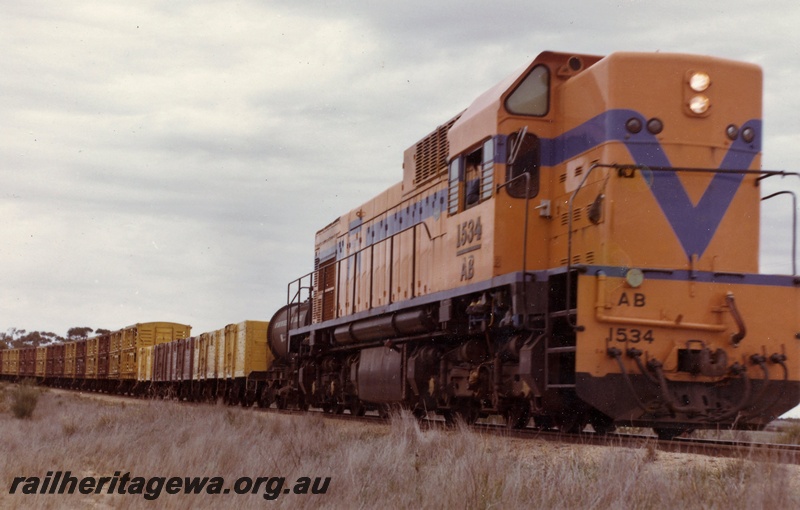 P15906
AB class 1534, in Westrail orange with blue stripe, on goods train comprising various wagons and vans, side and front view from track level
