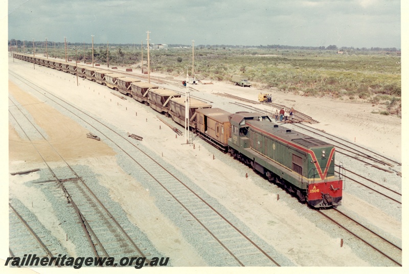 P15913
A class 1506, in green with red and yellow stripe, on goods train comprising brakevan and wagons, stopped at semaphore signal, track laying under way nearby, Kwinana, side and front view from elevated position
