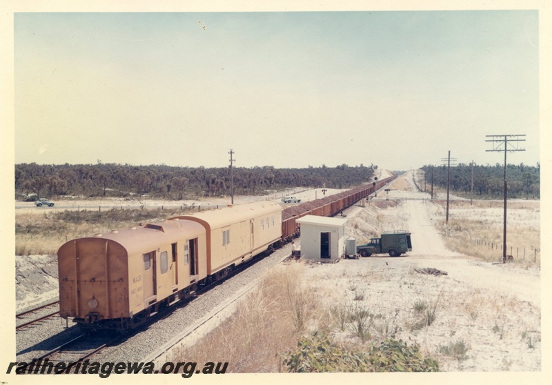 P15916
Iron ore train comprising vans and loaded wagons, level crossing, light signals, rural setting, trackside building, motor vehicles, rear and side view of train
