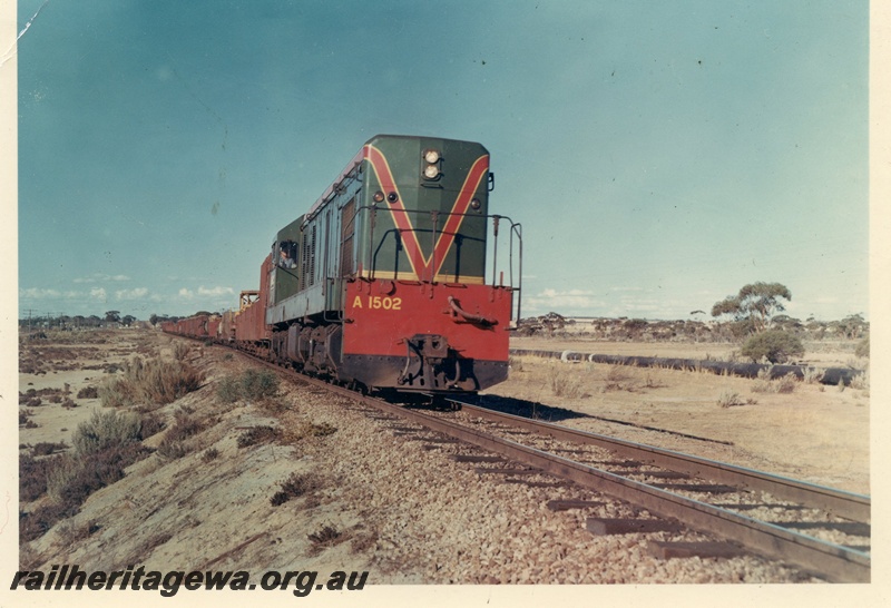 P15948
A class 1502, in green with red and yellow stripe, on goods train, rural setting, side and front view
