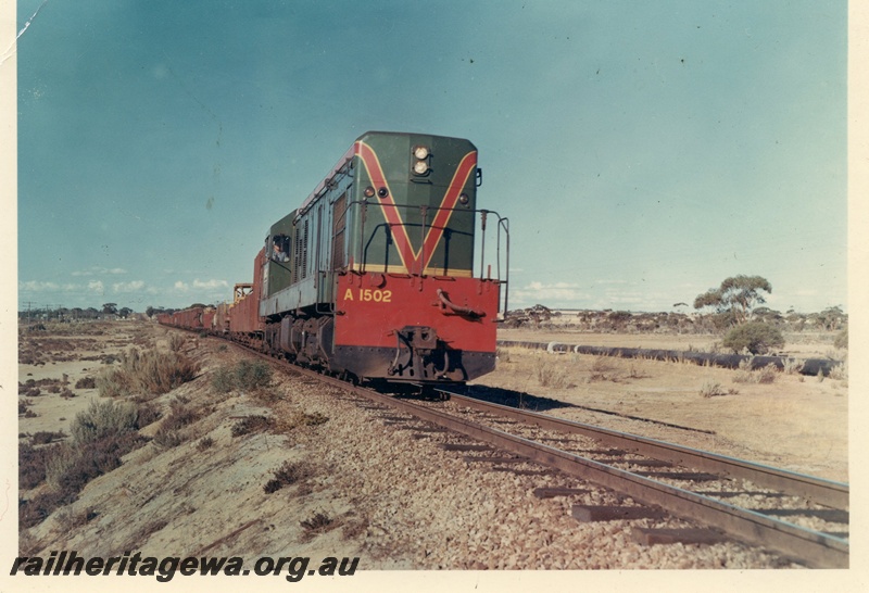 P15948
A class 1502, in green with red and yellow stripe, on goods train, rural setting, side and front view
