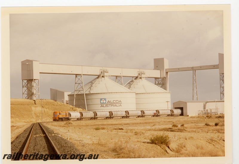 P15970
Alcoa Australia plant, conveyors, silos, RA class loco in Westrail orange with blue and white stripe, on train of hoppers partially in shed, distant view
