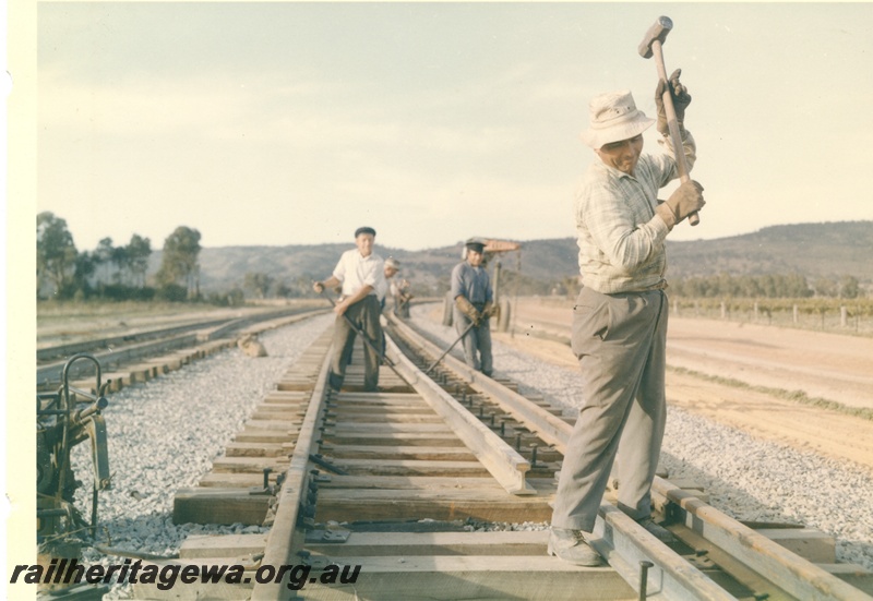 P16004
Track laying, worker driving spike into sleeper, other workers moving rail, rural setting, track level view
