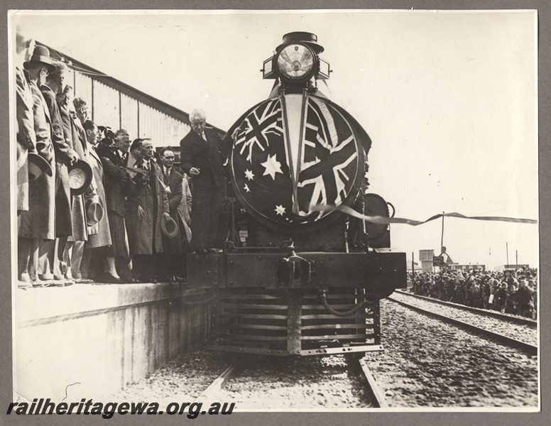 P16157
Commonwealth Railways (CR) G class 18, draped with British and Australian flags, on the official train at the opening of the Port Augusta to Port Pirie standard gauge extension of the TAR line, Prime Minister Joseph Lyons standing on the front of the engine, crowds of onlookers, front view
