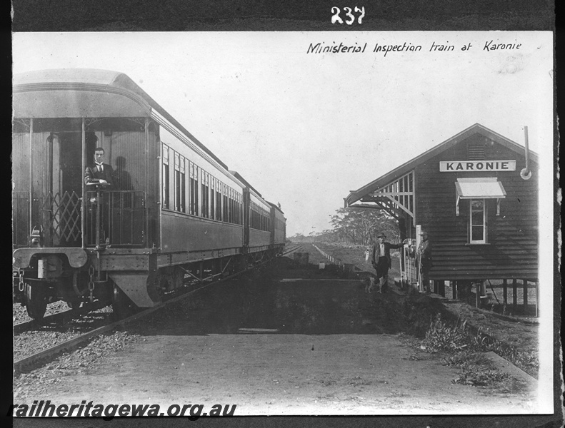P16163
Commonwealth Railways (CR), ministerial inspection train, rear to camera, suited man standing on outside platform of last carriage, station building on stilts, man and dog, Karonie, TAR line, view from rear of train
