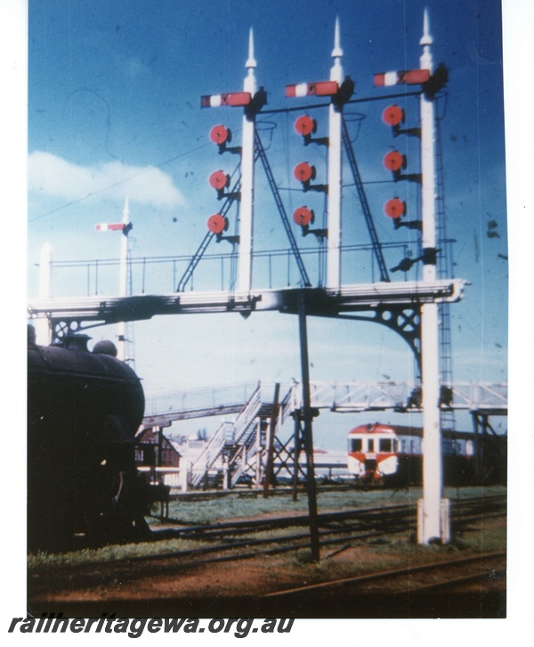 P16242
Signal gantry (part), DMU railcar set in red white and green livery, overhead footbridge, stair, North Fremantle, ER line
