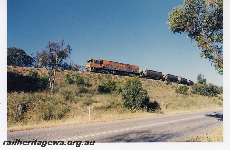 P16391
P class loco in Westrail orange with blue and white stripe, on goods train, on embankment near road, front and side view from road level 
