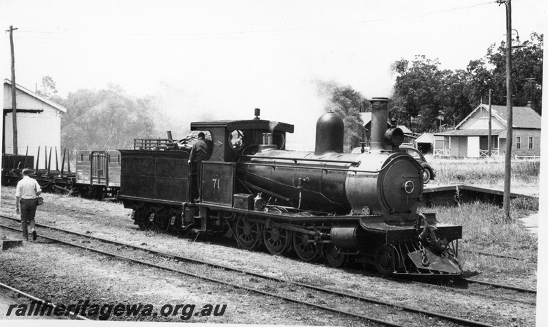 P17138
G class 71 locomotive reversing towards Vintage Train at Yarloop with portion of goods shed in background, open wagon & flat top wagon, on goods road, behind loco. Front & side view of loco. Note portion of goods loading ramp to right of loco and house in background. SWR line. 
