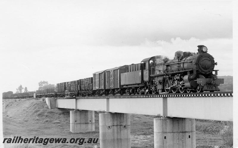 P17757
PMR class 734 steam locomotive hauling 23 goods train to Bunbury over the Roelands Bridge. SWR line. Steel girder concrete pylons on bridge. Side view of locomotive and train.
