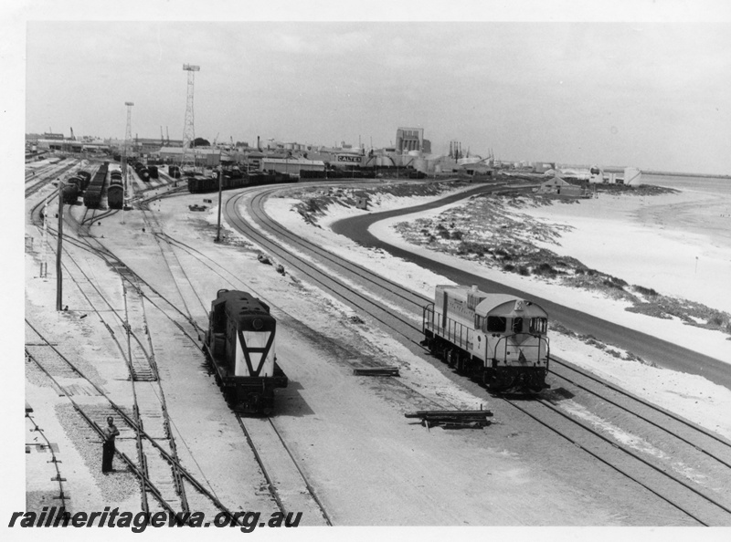 P17880
A general view of the western end of Leighton Yard showing unidentified J class standard gauge and Y class 1114 narrow gauge diesel locomotive. Photo taken from Yardmaster'ss Office block and tower lights can be seen in background as can the grain silos on North Wharf. 
