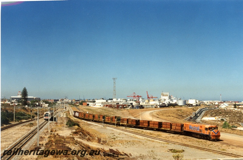 P17963
Overview of Leighton and Fremantle area, cranes and port buildings in background, suburban EMU from Fremantle and diesel hauled export jarrah timber train arriving Leighton yard in foreground, ER line
