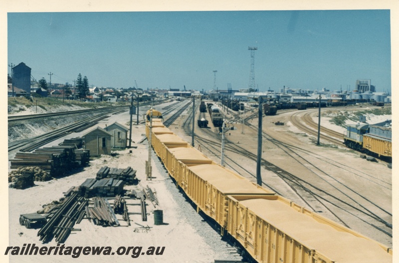 P17996
Rake of WG class wagons loaded with wheat, J class loco with WG class wagons modified to carry grain on the right hand side of the view, trackside buildings, sleepers, Leighton, Fremantle port in background, ER line, c1966
