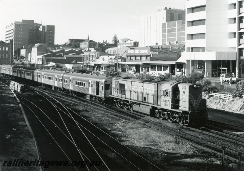 P18051
RA class 107, heading suburban passenger train comprising leased Queensland coaches, city buildings, arriving Perth City 
