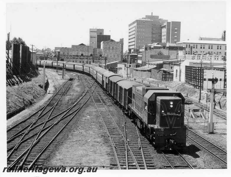 P18104
A class 1509 diesel locomotive hauling The Westland into Perth Station. ER line. Gantry signal frame over mid train, semaphore signal to right and trackage on left. Royal Perth Hospital is large building in background.
