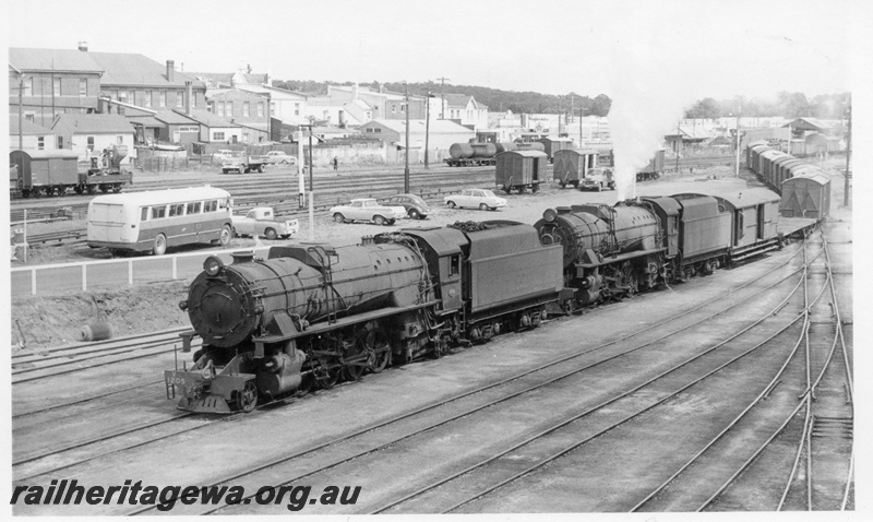 P18166
V class 1205 and V class 1222, double heading No 11 goods train into yard, carpark with bus and cars, vans, tanker cars, shops and buildings, Narrogin
