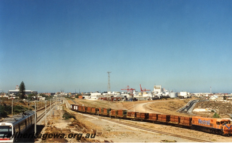 P18638
DB class diesel locomotive heading an export train load of jarrah. Similar to P18637. Full train length shown with background view of the fuel tanks at North Fremantle. A suburban Electric set is seen travelling to Perth at left of picture.
