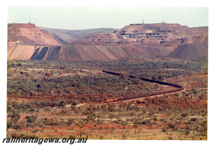 P18765
Mount Newman (MNM) a 192 car train departs Newman with Mount Whaleback mine in background. Photo shows the entire length of train hauled by 3 locomotives.
