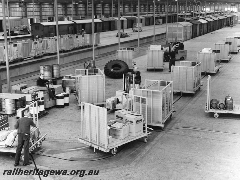 P20693
Interior view of outward shed of freight terminal, platforms, rakes of vans, Towveyor system in operation, trolleys, forklifts, workers, Kewdale, overview from elevated position 
