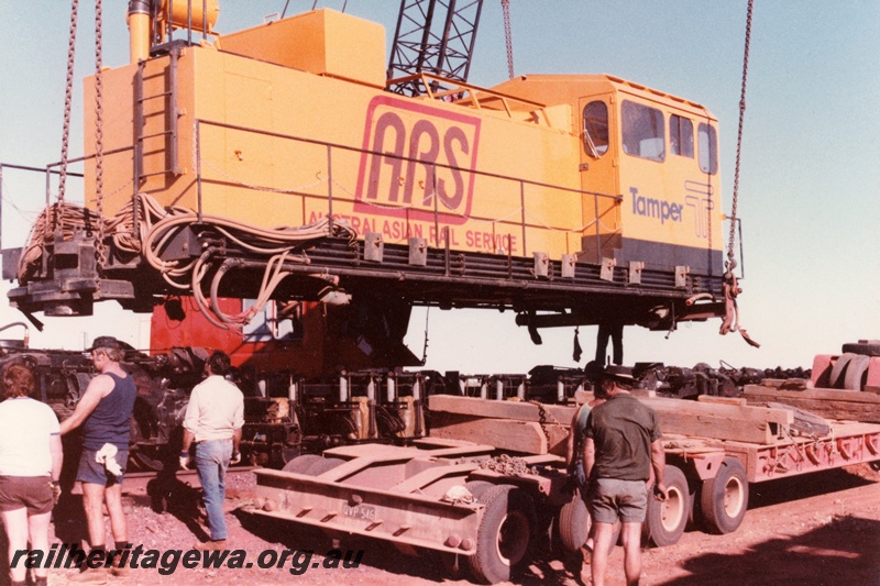 P21321
Loram Australian Rail Service rail grinder, middle unit being lowered by crane onto road trailer, workers, Port Hedland, end and side view from ground level 
