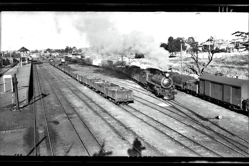 P21518
Steam loco on Perth bound goods train leaving yard, platform, signal box, rakes of wagons and vans, signals, signal shadows in foreground, houses in background, Northam, ER line, elevated view of western end of station and yard
