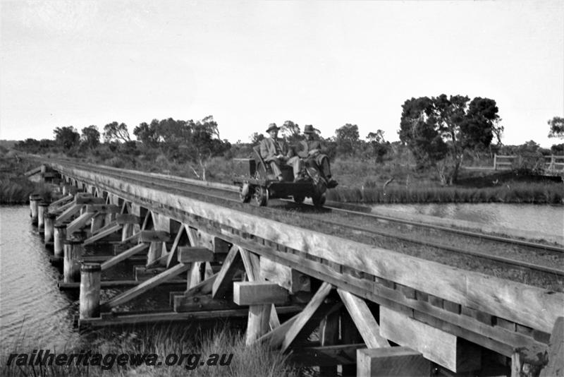 P21587
Rail trolley with two railwaymen on board, crossing wood trestle bridge, possibly Hay River Bridge, D line, side and front view from river bank 

