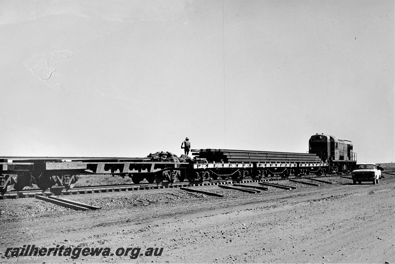 P22198
Construction of extension of Goldsworthy Mining Railway to tap deposits at Shay Gap, diesel loco on rail train, flat bed wagons loaded with rails, worker, motor vehicle, Pilbara, track level view 
