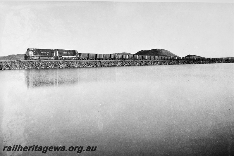 P22205
Hamersley Iron diesels numbered 2000 and 2015 double heading a train of empty wagons, on a rockfill causeway near Dampier, Pilbara, front and side view
