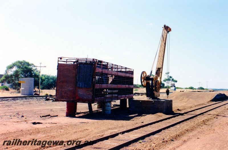 P23054
Stationary crane on concrete block, wheel-less stock wagon on drums, station shed with sign, tracks, Tardun, EM line, track level view looking south
