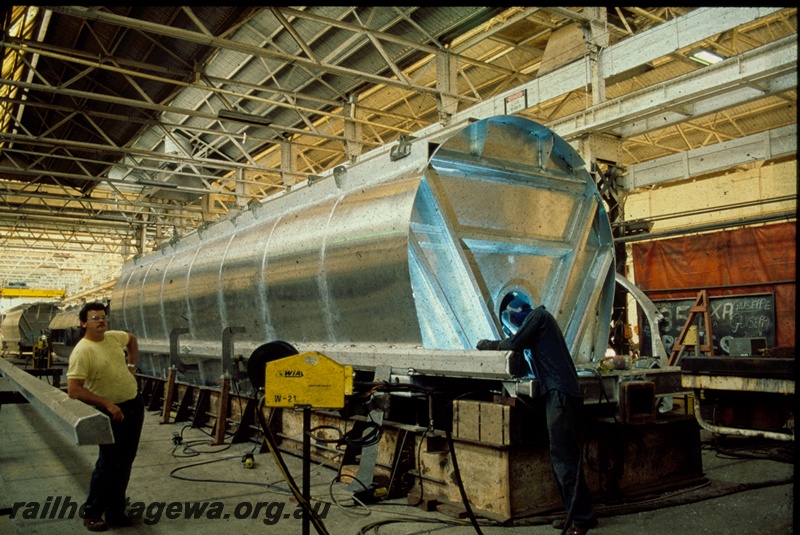 T00369
1 of 3 views of the construction of XU class wheat wagons, Boiler Shop South, Midland Workshops, wagon body on the main assembly jig, WAGR employees Bruce Skeggs (right) and Steve Butler (left) in the view.
