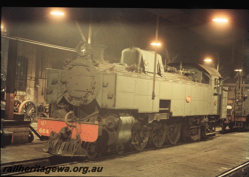 T04444
UT class 664 steam locomotive pictured inside the steam sheds at East Perth with the tender of a V class steam locomotive visible. Note the wooden flooring within the shed and overhead lights.
