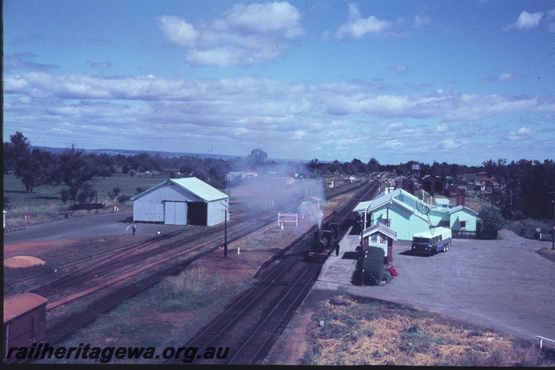 T04456
G class 118 steam locomotive, hauling another G class locomotive, departing Pinjarra enroute to Forrestfield for disposal. Overhead view of Station buildings and goods shed. Note WAGR passenger/freighter bus in parking area. SWR line.
